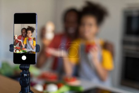 Téléchargez les photos : Blog de famille. Père noir et fille préadolescente capturant la vidéo tout en cuisinant dans la cuisine, père afro-américain et enfant féminin préparant des aliments sains à la caméra, foyer sélectif à l'écran du téléphone intelligent - en image libre de droit