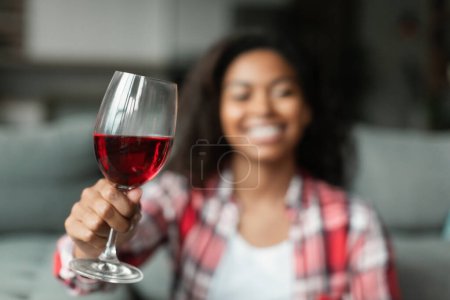 Foto de Sonriente milenaria dama negra aplaude copa de vino a la cámara en el interior de la sala de estar, borrosa. Celebración navideña, felicitaciones y fechas remotas, descanso y relax con alcohol, nueva normalidad en casa - Imagen libre de derechos