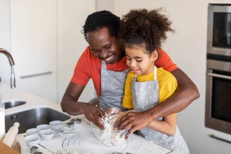 Téléchargez les photos : Prendre soin du papa afro-américain enseignant à sa fille comment faire de la pâte pendant qu'ils cuisinent ensemble à la maison, jeune père noir et préadolescente enfant féminin préparant la pâtisserie dans la cuisine, espace libre - en image libre de droit