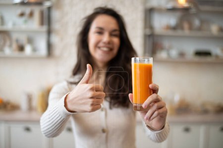 Foto de Señora caucásica positiva sosteniendo vaso de jugo de naranja orgánico y mostrando el pulgar hacia arriba, de pie en el interior de la cocina de luz. Nutrición adecuada, desayuno, concepto de atención médica - Imagen libre de derechos