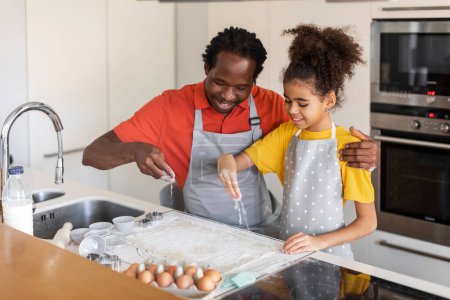 Foto de Padre negro sonriente e hija haciendo galletas juntos en la cocina, feliz papá afroamericano y preadolescente niña usando delantales horneando en casa, preparando la masa para la pastelería, espacio libre - Imagen libre de derechos