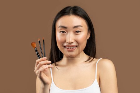 Téléchargez les photos : Concept de maquillage. Portrait de jeune femme coréenne tenant un ensemble de pinceaux de maquillage, debout sur fond de studio brun et souriant à la caméra, espace de copie - en image libre de droit