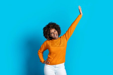 Photo for Joyful happy beautiful african american young woman with bushy hair in stylish casual outfit moving on blue studio background, raising one hand up and smiling, black lady dancing alone - Royalty Free Image