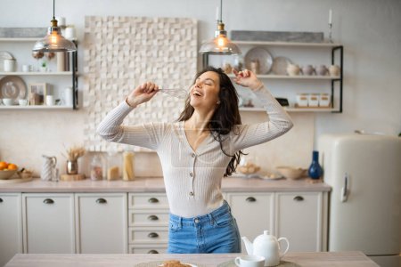Photo for Playful young woman singing at appliance as imaginary microphone, dancing in light kitchen interior, copy space. Fun at home alone, lady superstar enjoy free time - Royalty Free Image