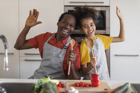 Téléchargez les photos : Joyeux père noir et fille de préadolescence s'amusant tout en cuisinant ensemble dans la cuisine, heureux père afro-américain et enfant féminin chantant et dupant, en utilisant des cuillères à spatule comme microphones - en image libre de droit