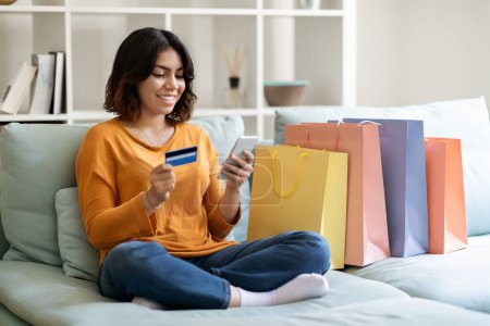 Photo for Online Shopping Concept. Happy Arab Female Using Smartphone And Credit Card While Sitting On Couch With Bright Shopper Bags At Home, Smiling Middle Eastern Woman Making Internet Purchases, Free Space - Royalty Free Image