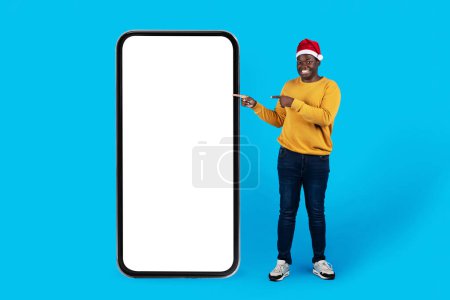 Photo for Online Ad. Cheerful Black Male In Santa Hat Pointing At Big Blank Smartphone Screen While Standing Over Blue Studio Background, Happy Young African Man Recommending New Mobile App, Collage, Mockup - Royalty Free Image