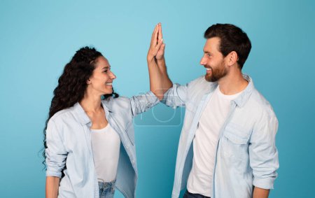 Photo for Smiling young european man and arab lady make high five hands sign, celebrating victory and success, have fun, isolated on blue background, studio, copy space. Emotions from win, good ad and offer - Royalty Free Image