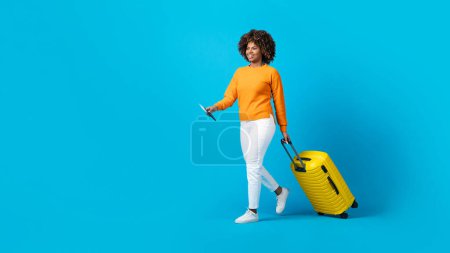 Téléchargez les photos : Femme afro-américaine millénaire excitée voyageant seule, portant des bagages jaunes, tenant un passeport avec des billets d'avion, marchant sur fond bleu studio, espace de copie, pleine longueur, bannière - en image libre de droit