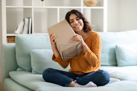 Photo for Portrait Of Happy Young Arab Woman Embracing Cardboard Box With Delivery At Home, Smiling Middle Eastern Female Sitting On Couch With Delivered Parcel In Hands, Enjoying Fast Shipping Services - Royalty Free Image