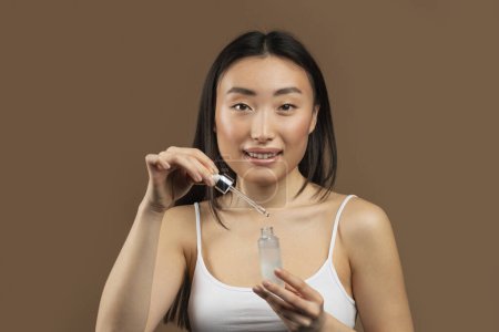 Téléchargez les photos : Heureuse jeune femme asiatique utilisant du sérum ou de l'huile dans sa routine quotidienne de soins du visage, recommandant de nouveaux produits de beauté, posant sur fond de studio brun - en image libre de droit