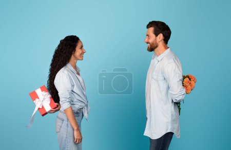 Photo for Smiling handsome young european husband and arab wife hold gift and flowers behind back, isolated on blue background, studio. Birthday, holiday greeting, anniversary gift and valentine day celebrate - Royalty Free Image