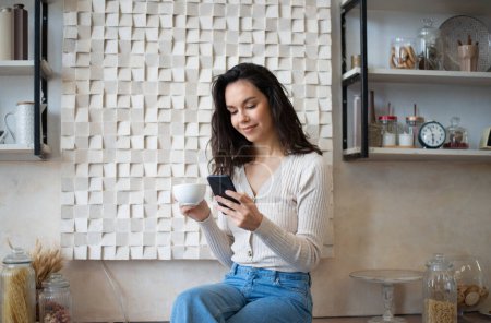 Photo for Positive lady drinking hot morning drink and chatting on smartphone, reading message, sitting in kitchen interior. Social networks, blog and new app, ad and good offer - Royalty Free Image