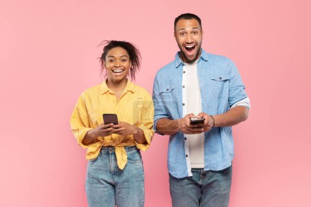 Photo for Mobile offer. Portrait of overjoyed black couple holding smartphones and looking at camera with excitement, excited man and woman opening mouth with amazement, posing over pink background - Royalty Free Image