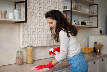 Photo for Excited housewife in rubber gloves cleaning counter with spray detergent at kitchen, free space. Young maid keeping her flat tidy, doing domestic chores, disinfecting apartment - Royalty Free Image