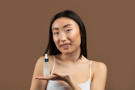 Foto de Concepto de cuidado facial. Mujer asiática mostrando botella de suero facial, posando sobre fondo de estudio marrón. Mujer coreana publicidad cosméticos favoritos y sonriendo a la cámara - Imagen libre de derechos