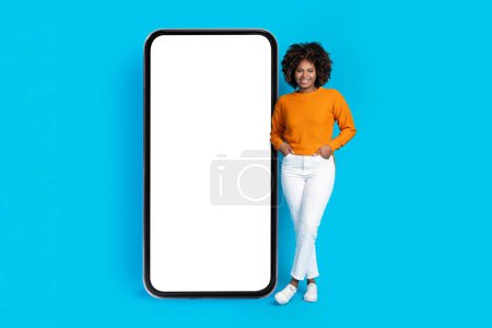 Photo for Cool attractive african american young lady with bushy hair in nice stylish outfit posing with hands in pants pockets by big phone with white empty screen, mockup, blue background, full length - Royalty Free Image