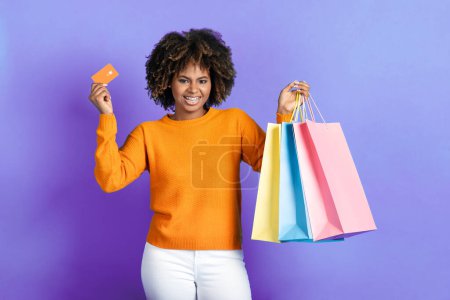Photo for Positive stylish young african american lady with bushy hair and braces posing with credit card and purchases colorful shopping bags over purple studio background. Retail, shopping concept - Royalty Free Image