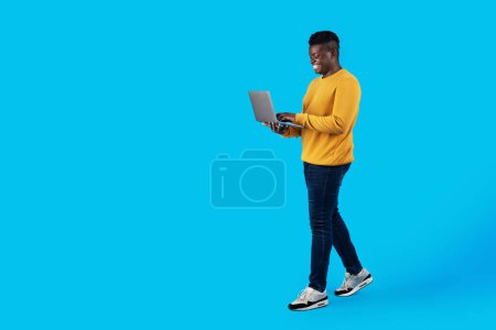 Photo for Cheerful Young Black Man With Laptop Walking Over Blue Studio Background, Full Length Shot Of Millennial African American Guy Using Computer For Remote Work Or Online Shopping, Copy Space - Royalty Free Image