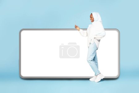 Photo for Excited black woman in hijab standing near big cellphone with empty screen and using mobile device on blue studio background, mock up for website or application design - Royalty Free Image