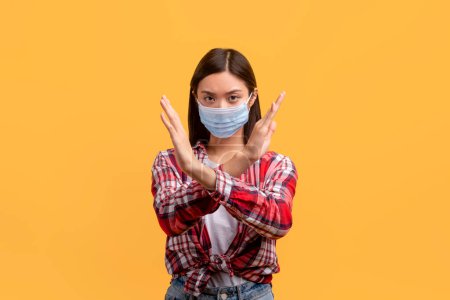 Foto de Mujer asiática usando mascarilla médica y mostrando gesto de alto con los brazos cruzados, protestando por algo, diciendo no a la cuarentena del coronavirus, moviéndose sobre el fondo amarillo - Imagen libre de derechos