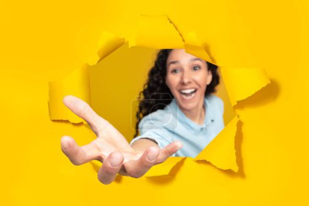 Téléchargez les photos : Jeune femme excitée étirant la paume ouverte à travers un trou dans du papier jaune déchiré, prenant ou donnant quelque chose, tenant un objet invisible et souriant, espace de copie - en image libre de droit