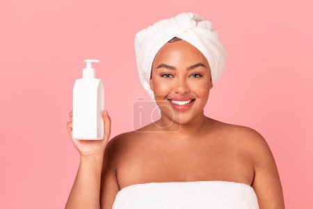 Photo for Attractive african american plus size lady with bottle of moisturising body lotion, posing over pink background. Beautiful woman standing wrapped in towel, enjoying new beauty product - Royalty Free Image