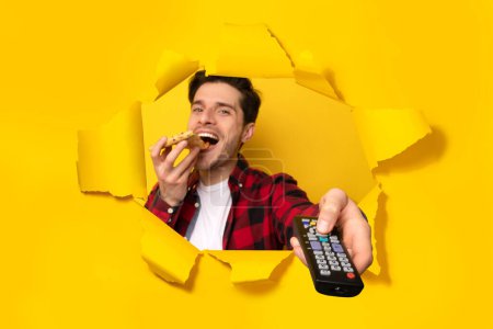 Foto de Hombre viendo la televisión, apuntando mando a distancia y comiendo pizza, posando en un agujero de papel amarillo roto. Programación televisiva, tecnología y entretenimiento. Collage. - Imagen libre de derechos