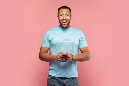 Photo for Handsome surprised black man holding smartphone and looking at camera in shock on pink studio background. Emotional guy with mobile phone amazed at promo sale or huge discount - Royalty Free Image