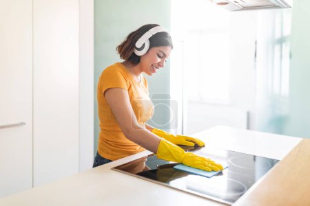 Photo for Happy Middle Eastern Female Listening Music In Headphones While Cleaning Kitchen, Cheerful Young Arab Woman Polishing Induction Cooktop Surface With Rag And Enjoying Favorite Songs, Copy Space - Royalty Free Image
