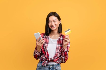 Photo for Easy money transfer. Happy young asian lady holding debit credit card and using mobile phone, making online payment standing isolated on yellow studio background - Royalty Free Image