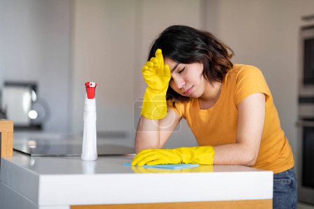 Mujer árabe joven sintiéndose cansada mientras hace la limpieza en la cocina en casa, mujer de Oriente Medio que usa guantes de goma agotados después de lavar la mesa, tener descanso y limpiar la frente, espacio libre