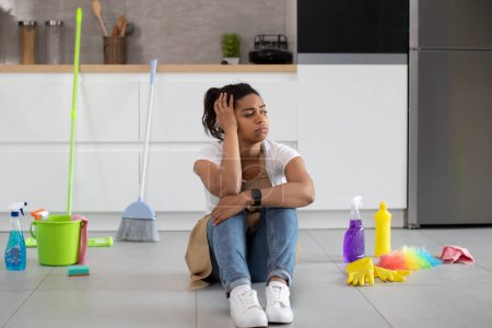Photo for Upset tired millennial african american housewife in apron sits on floor with cleaning supplies, looks at mess in dirty kitchen interior. Chaos and problems with cleaning, break from many work at home - Royalty Free Image