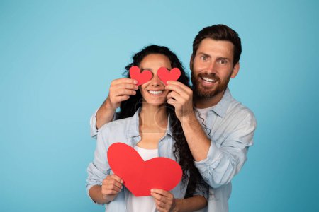 Photo for Glad young european husband in casual clothes puts hearts on arab wife eyes, have fun, isolated on blue background, studio. Family love and relationships, valentine day and anniversary celebration - Royalty Free Image