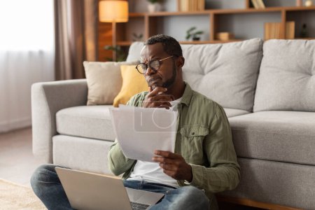 Photo for Pensive serious adult black man in glasses uses computer, works with documents, sits on floor in living room interior. Data analysis, gadget for business and mistake in work remotely, startup at home - Royalty Free Image