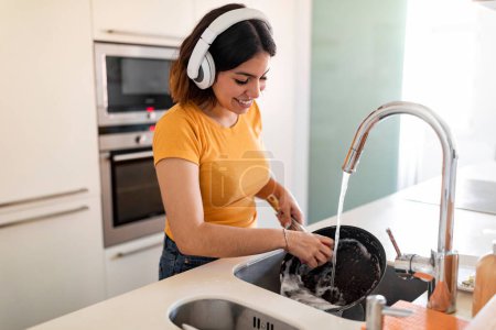 Foto de Sonriente Oriente Medio Femenino con auriculares inalámbricos haciendo platos en la cocina, Feliz joven ama de casa árabe Lavadora con esponja y detergente y disfrutando de música favorita, espacio libre - Imagen libre de derechos