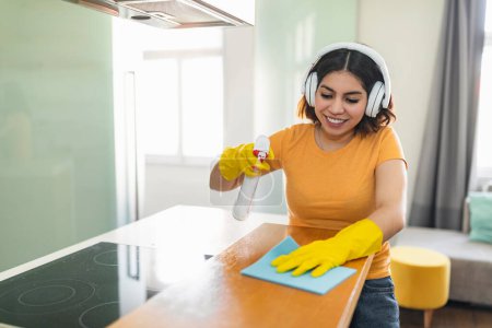 Photo for Happy Middle Eastern Female Cleaning Kitchen Counter With Detergent Spray And Rag, Smiling Young Arab Woman Washing Surface And Listening Music In Wireless Headphones, Enjoying Home Chores - Royalty Free Image