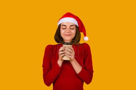 Photo for Portrait of attractive cheerful dreamy brunette young woman wearing Santa hat drinking smelling tasty cacao with closed eyes, isolated over orange color studio background, copy space - Royalty Free Image