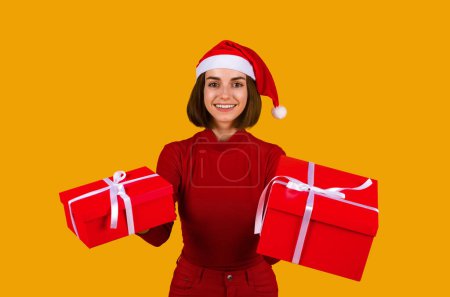 Foto de Linda mujer morena bonita sonriente en sombrero rojo Santa Claus sosteniendo regalos rojos de Navidad, feliz saludo dama con Navidad y Año Nuevo 2023 sobre fondo de estudio naranja, espacio de copia - Imagen libre de derechos