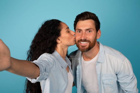 Téléchargez les photos : Jeune femme européenne souriante en baisers décontractés homme barbu et prend selfie, profiter d'un moment tendre isolé sur fond bleu, studio, gros plan. Amour et relation, photo pour blog et réseaux sociaux - en image libre de droit