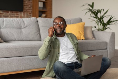 Foto de Feliz adulto sonriente hombre de negocios negro en gafas utiliza la computadora, llamadas por teléfono, gesticula, se sienta en el suelo en el interior de la sala de estar, espacio libre. Dispositivo para negocios, trabajo, comunicación remota en casa - Imagen libre de derechos