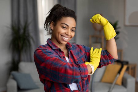 Foto de Glad millennial mujer afroamericana en guantes de goma muestra signo de fuerza con la mano y listo para las tareas domésticas en el interior de la sala de estar. Gesto para un excelente servicio de limpieza, muchos trabajan en casa - Imagen libre de derechos