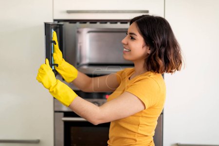 Photo for Housekeeping Concept. Smiling Arab Woman In Rubber Gloves Wiping Microwave From Dirt While Doing Cleaning In Kitchen, Happy Young Middle Eastern Housewife Tidying Home, Side View, Closeup Shot - Royalty Free Image