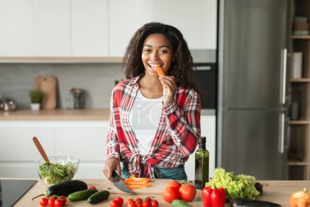 Photo for Smiling pretty millennial black lady preparing salad, eating carrot at table with organic vegetables in kitchen interior, free space. Health care, vegan food, prepare lunch, proper nutrition at home - Royalty Free Image