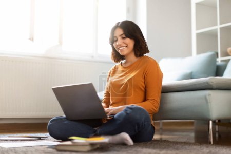 Téléchargez les photos : Enseignement à distance. Joyeux jeune Moyen-Orient étude féminine avec ordinateur portable à la maison, femme arabe gaie assise sur le sol avec ordinateur dans le salon, bénéficiant d'une éducation en ligne, Copier l'espace - en image libre de droit