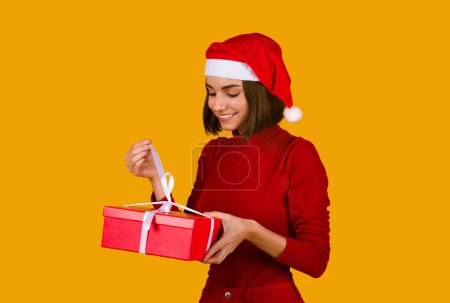 Téléchargez les photos : Joyeux excité belle jeune femme dans santa claus chapeau avec coiffure mignonne tenant boîte cadeau rouge sur fond de studio orange, cadeau d'ouverture de Noël, Noël et Nouvel An 2023 fête - en image libre de droit