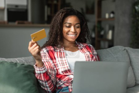 Photo for New buy and app for pay. Happy millennial black lady using computer and credit card to check bank account, sitting on sofa in living room interior. Online shopping, big sale and cashback and home - Royalty Free Image