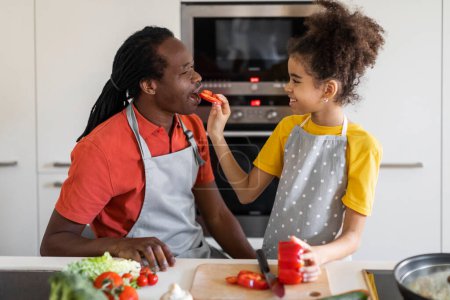 Foto de Retrato del amoroso padre negro y su hija preparando comida en la cocina juntos, sonriendo preadolescente niña alimentando a su padre feliz con rodaja de pimiento mientras hacen ensalada de verduras en casa - Imagen libre de derechos