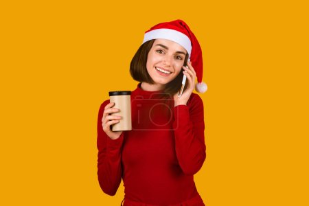 Téléchargez les photos : Jolie dame brune positive au chapeau de Père Noël et tenue rouge parlant sur téléphone portable et souriant, tenant une tasse en papier, buvant du café chaud, du thé ou du cacao, isolée sur fond de studio orange, espace de copie - en image libre de droit
