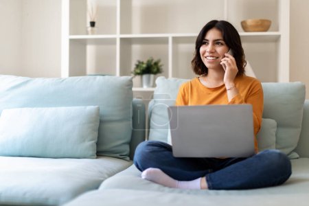 Photo for Remote Business. Smiling Arab Woman Talking On Phone And Using Laptop At Home, Beautiful Young Middle Eastern Female Sitting On Couch In Living Room, Working On Computer And Having Mobile Call - Royalty Free Image